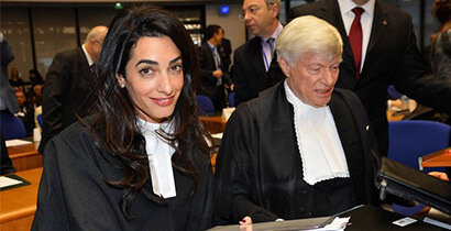Two professionals in courtroom robes sit at a desk; the woman smiles at the camera beside an older man reviewing documents.