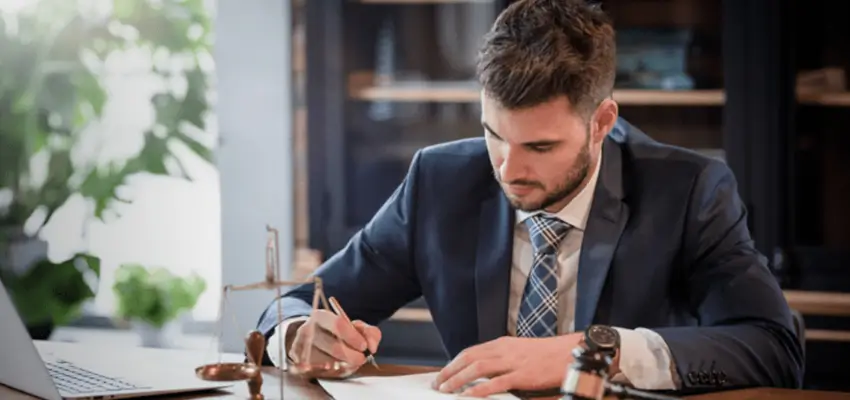Professional man in a suit signing papers at a desk, with a gavel and scales nearby in a bright office setting