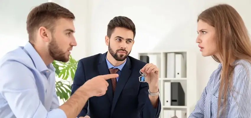 Three coworkers in a bright office: a man in a suit holds up a keyring as another man points and a woman looks on attentively ground-level discussion about keys and an item being shown