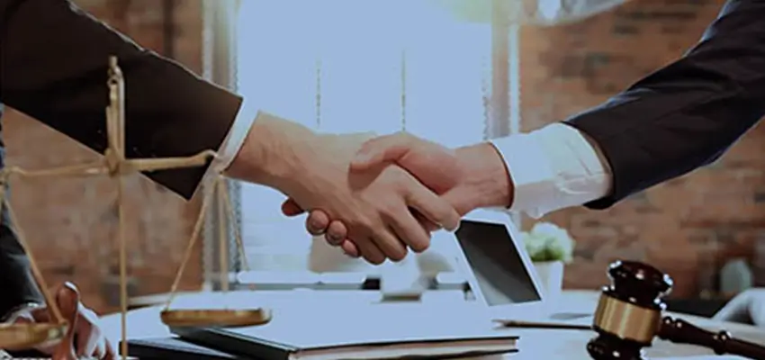 Two professionals shake hands over a desk, with legal symbols like a gavel and scales in the background, suggesting a deal or agreement.