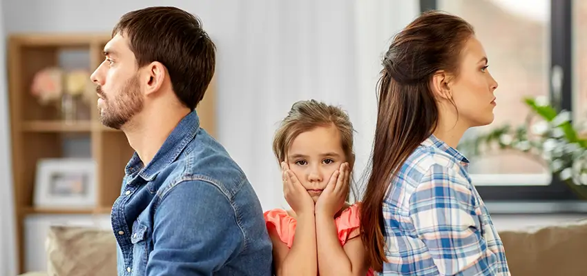 Parents sit back-to-back on a sofa with a young girl between them, looking distressed as she rests her face in her hands.