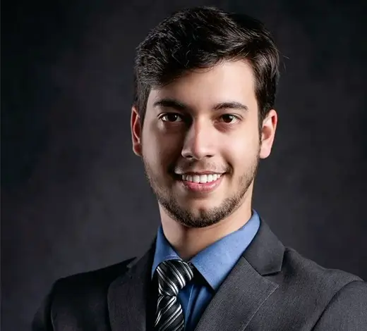 Young man in a dark suit and blue shirt, smiling for a professional headshot against a dark studio background.
