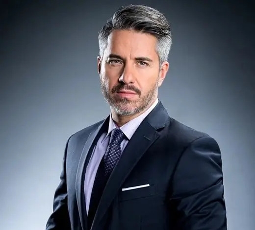 Professional headshot of a middle-aged man in a navy suit, light purple shirt, and dark tie, posing against a gray gradient background.